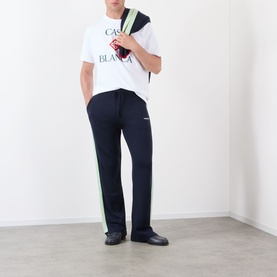 Model wearing Casablanca Diamond Logo T-Shirt in white with stacked logo print, styled with navy track pants and green crossbody bag.