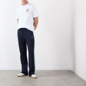 Model wearing Casablanca Classic Logo Graphic T-Shirt in white with chest graphic, styled with navy track pants with green side stripe.