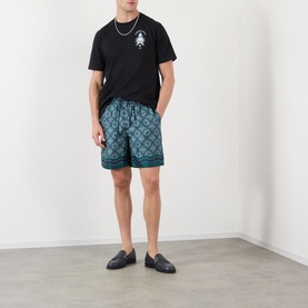 Model wearing Casablanca Radiating Monogram silk shorts in teal, styled with black T-shirt and dark loafers, hands in pockets.