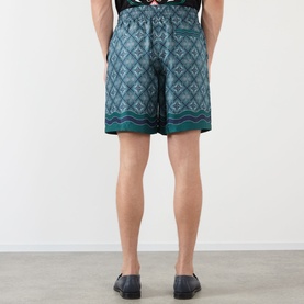 Model wearing Casablanca Shorts in Radiating Monogram print, teal silk with geometric pattern, rear view, styled with navy loafers