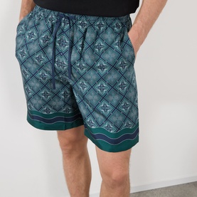 Model wearing Casablanca silk shorts in Radiating Monogram print, teal geometric pattern with contrast border hem, elasticated waist
