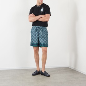 Model wearing Casablanca Radiating Monogram silk shorts in teal geometric print, styled with black T-shirt and navy loafers