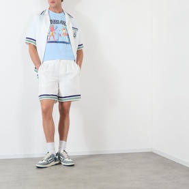 Model wearing Casablanca Egyptian Tennis Match Logo T-Shirt in light blue, styled with white stripe-trim shorts and grey trainers.