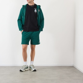 Model wearing Casablanca Hotel Emblem black cotton T-shirt, styled with green zip hoodie, green shorts, and grey trainers.