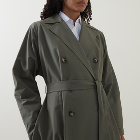 Model wearing olive green double-breasted belted trench coat with tortoiseshell buttons, styled over a white shirt