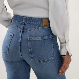 Model wearing Hugo Boss medium blue high-waist slim jeans, rear view showing back pockets and leather logo patch, styled with white blouse.