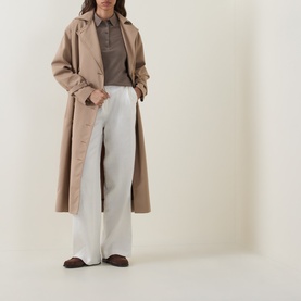Model wearing Hugo Boss Fidenka Knitted Polo Shirt in Brown Overflow, styled with a beige trench coat, wide-leg white trousers and brown loafers.