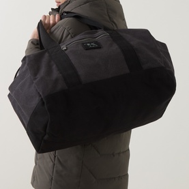 Model carrying Rodd & Gunn Foxton Duffle Holdall in Charcoal Onyx, denim finish with contrast logo, zip pocket and gold-tone hardware visible.
