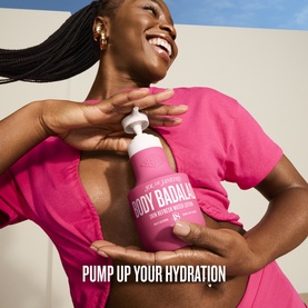 Model wearing pink dress, pumping Sol De Janeiro Body Badal&aacute; hydrating body lotion from a large pink bottle, smiling.