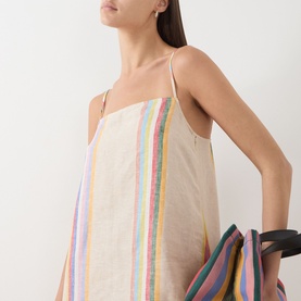 Model wearing Staud Day Trip Stripe linen dress with thin straps, styled with a multicoloured striped tote bag.
