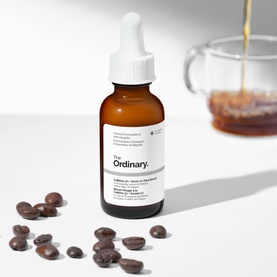 The Ordinary serum in amber dropper bottle, surrounded by coffee beans with a glass of coffee in the background.