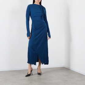 Model wearing teal blue long-sleeve midi dress with draped bodice and pleated fringe hem, styled with black pointed heels