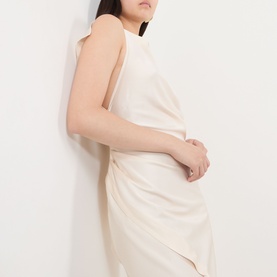 Model wearing a sleeveless cream midi dress with draped skirt detail and open back, leaning against a white wall