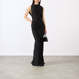 Model wearing Victoria Beckham Kyra Draped Gown in black, sleeveless with gathered side detail and floor-sweeping hem, styled with a small black bag