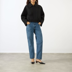 Model wearing Victoria Beckham black cropped jacket with elasticated hem, styled with blue straight-leg jeans and black kitten heel mules.