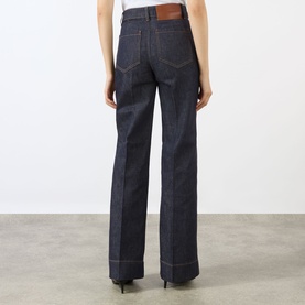 Model wearing Victoria Beckham Alina High-Rise Flared Leg Jeans in indigo, rear view showing leather patch, tobacco stitching and wide-leg silhouette