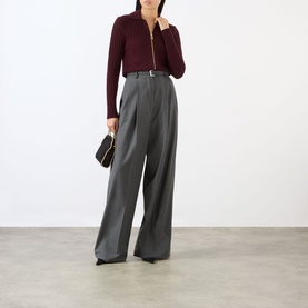 Model wearing Victoria Beckham Zip Knitted Polo Sweater in Acai with wide-leg grey trousers, styled with a black belt and clutch bag.
