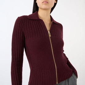 Model wearing Victoria Beckham Zip Knitted Polo Sweater in Acai, featuring ribbed construction and gold-tone zip, styled with grey trousers