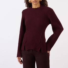 Model wearing burgundy ribbed knit crew neck sweater with flared cuffs and asymmetric hem, styled with dark trousers
