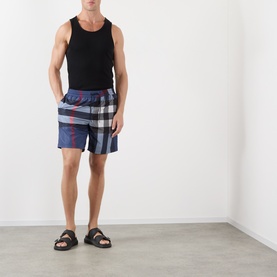 Model wearing Burberry carbon blue check swim shorts with black vest top and black sandals.