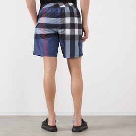 Model wearing Burberry carbon blue check swim shorts, rear view, paired with black slip-on shoes.