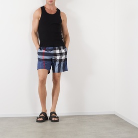 Model wearing Burberry carbon blue check swim shorts, styled with a black vest top and brown sandals.