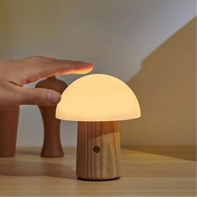 Small glowing mushroom-shaped night light with warm white silicone cap and wooden stem base, hand shown touching the top to activate