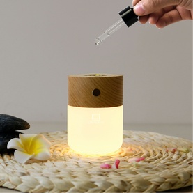 Cylindrical wood-effect aroma diffuser with glowing warm light base, shown with dropper adding essential oil, styled with flowers and stones