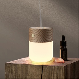 Cylindrical ultrasonic aroma diffuser with wood-grain top and glowing white base, emitting mist, styled with an essential oil bottle on a wooden