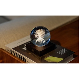 Gingko crystal ball light with engraved tree design on dark wooden base, displayed on a coffee table book in warm ambient lighting.