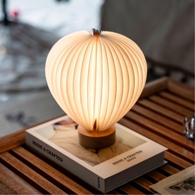 Gingko hot air balloon-shaped ribbed table lamp with warm glow, wooden base, displayed on a hardback book on a slatted wooden surface
