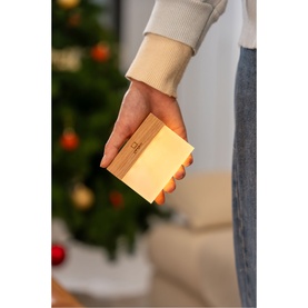 Hand holding a small glowing wooden cube lamp with warm light, Christmas tree visible in background