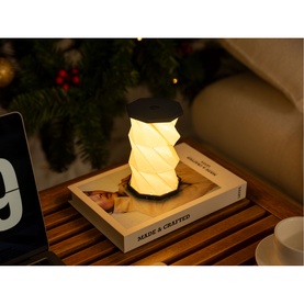 Gingko Mini Slate Black lamp glowing warm white, twisted column design, displayed on books atop a wooden surface with festive decor