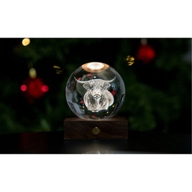 Gingko crystal ball light with 3D engraved highland cow motif, on a walnut wood base, glowing warmly against a festive background.