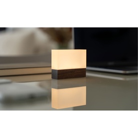 Small square ambient lamp with warm white glow and dark wood base, placed on a reflective surface with laptop in background
