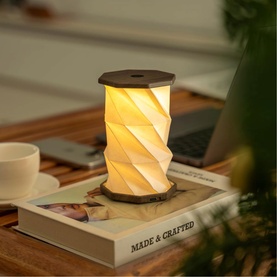 Gingko Mini Walnut Drift light glowing warm white with geometric spiral shade, placed on a book on a wooden desk