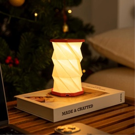 Gingko Mini Coral Red portable lamp with glowing twisted shade, styled on a book near a Christmas tree in a warm home setting