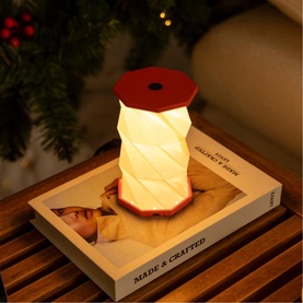 Gingko Mini Coral Red table lamp with geometric twisted body, warm glowing shade, styled on a book beside festive greenery