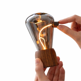 Gingko Edison bulb lamp with warm filament glow, held in hands above a walnut wooden base, being switched on