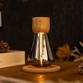 Smoked glass table lamp with wooden top and cork coaster base, glowing warmly on a wooden surface with pine cones in background