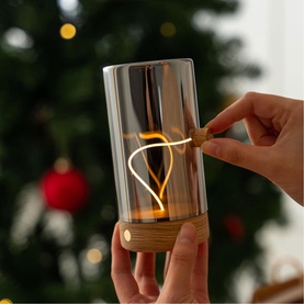 Cylindrical glass lantern with warm LED filament bulb and wooden base, switched on by hand, styled near a Christmas tree