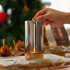 Silver glass and wood cordless table lamp being lit by hand, styled with festive Christmas tree backdrop and dried botanicals