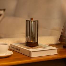 Gingko cylindrical table lamp with smoked glass shade and walnut wood base, glowing warmly on a bedside book stack