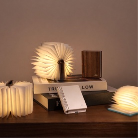 Gingko Smart Book Light in Walnut Finish, shown open and glowing alongside other sizes and finishes on a wooden surface.