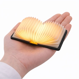 Gingko Space Black folding book lamp held in one hand, glowing warm amber light between fanned pages, compact size
