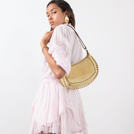 Model carrying Isabel Marant Oksan Moon Crossbody Bag in straw with gold stud trim detailing, styled with a pink tiered mini dress and hoop earrings