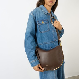 Model carrying Isabel Marant Oskan Soft Zip Crossbody Bag in coffee leather with gold studs and hardware, worn crossbody over denim jacket