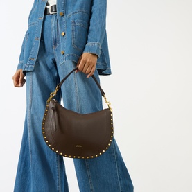 Model carrying Isabel Marant Oskan Soft Zip Crossbody Bag in coffee leather with gold stud embellishment and short top handle, styled with denim