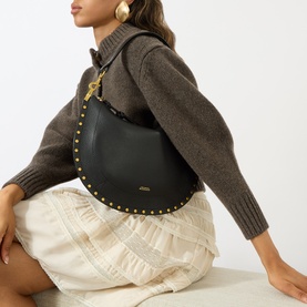 Model carrying Isabel Marant Oskan Soft Zip Crossbody Bag in black leather with gold studs and hardware, styled with brown knit jumper and cream skirt