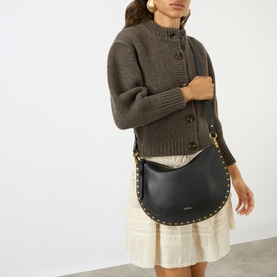 Model carrying Isabel Marant Oskan Soft Zip Crossbody Bag in black leather with gold studs and hardware, styled with brown knit and cream skirt